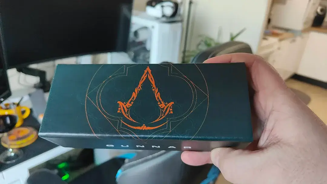 Close up photo of the Intercept Assassin's Creed Mirage edition glasses box. The box is square shaped with a sandy gold color and features the iconic Assassin's Creed logo.