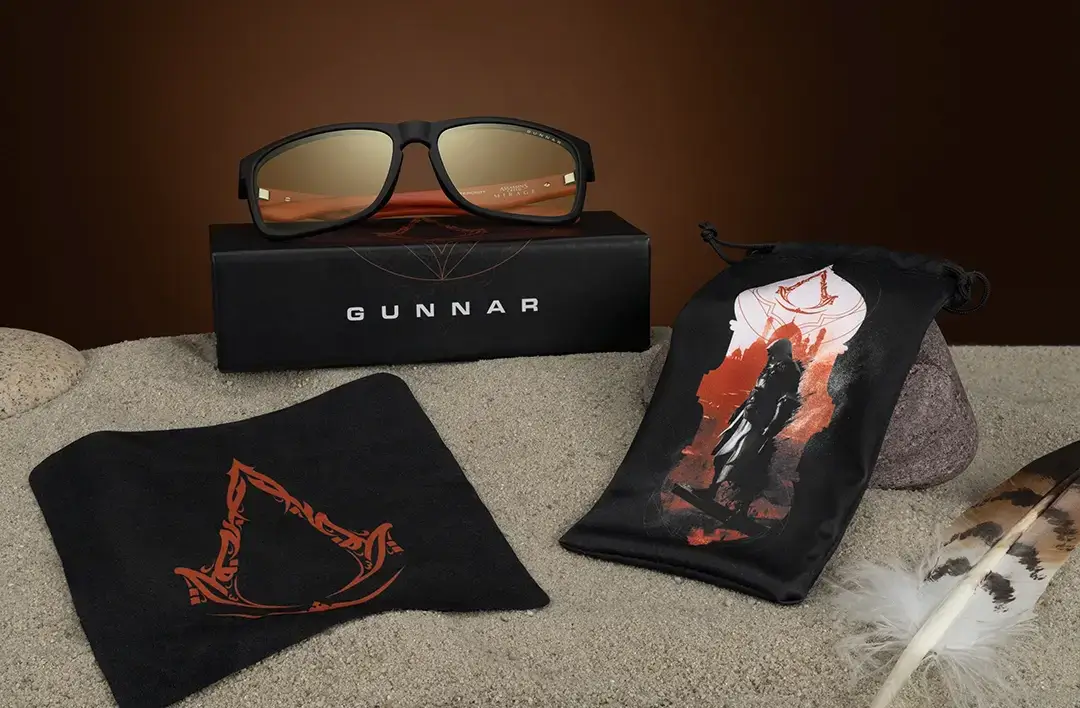 An overhead product shot showcasing the Intercept Assassin's Creed Mirage glasses contents including the glasses, box, pouch bag, and microfiber cloth all bearing the iconic Assassin's Creed logo.