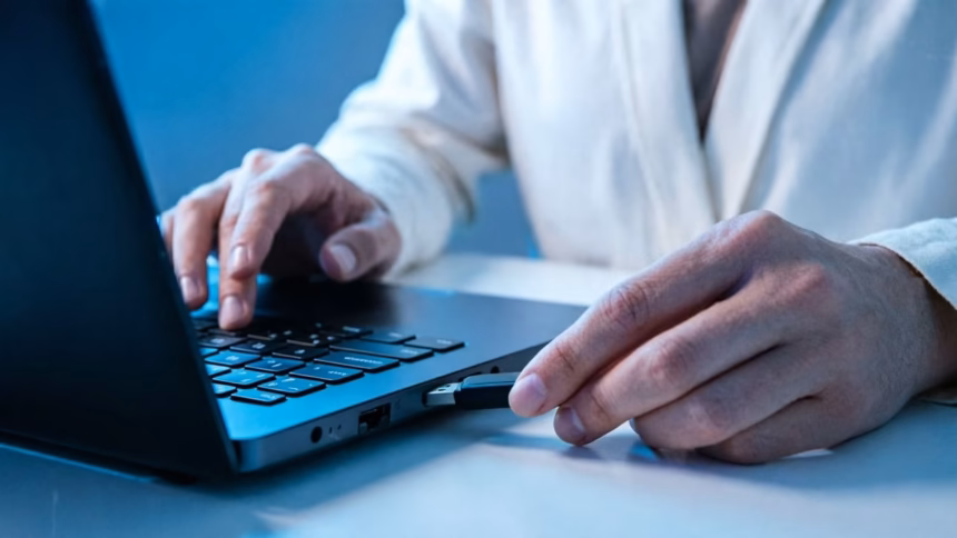 The $10 Keychain Gadget That Could Save Your PC | Smart Tech, Sharp Takes - CRYOVEX.com A small silver keychain usb flash drive attached to a metal ring, resting on a dark desk surface near a laptop keyboard.